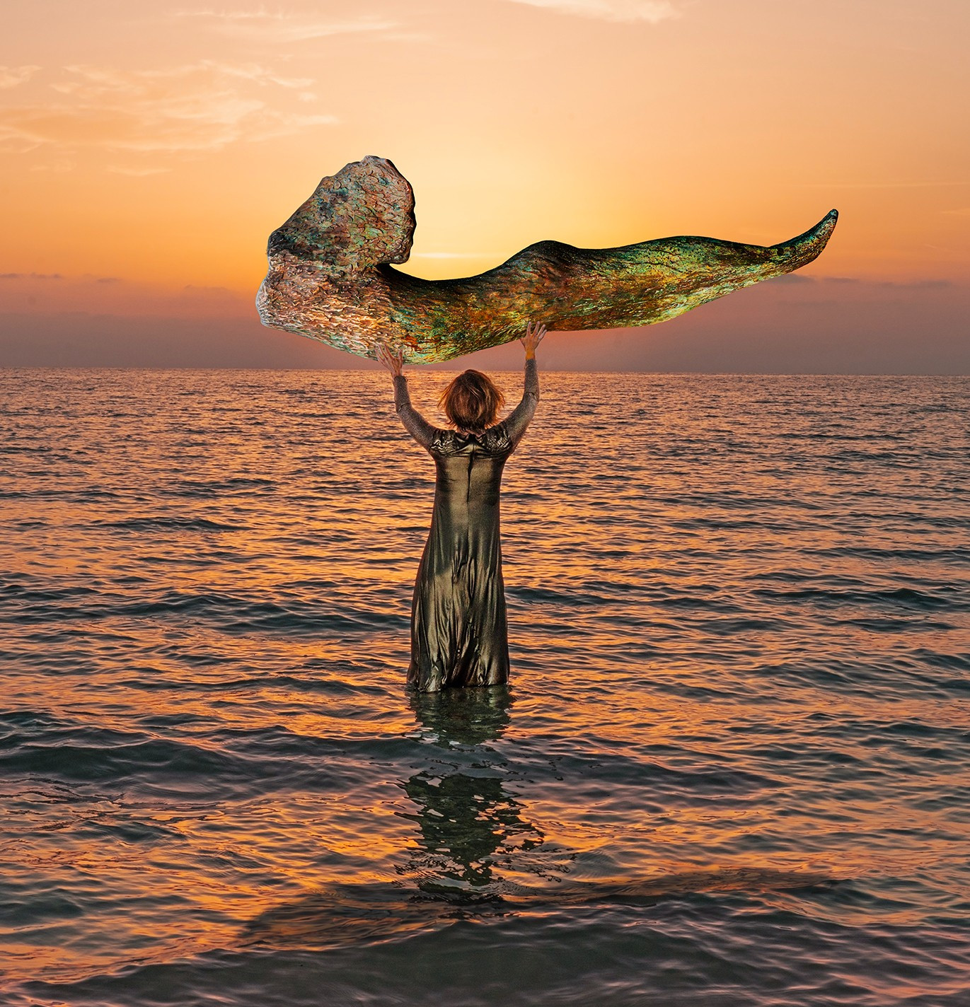 woman standing in ocean at sunset holding above her head a metal sculpture