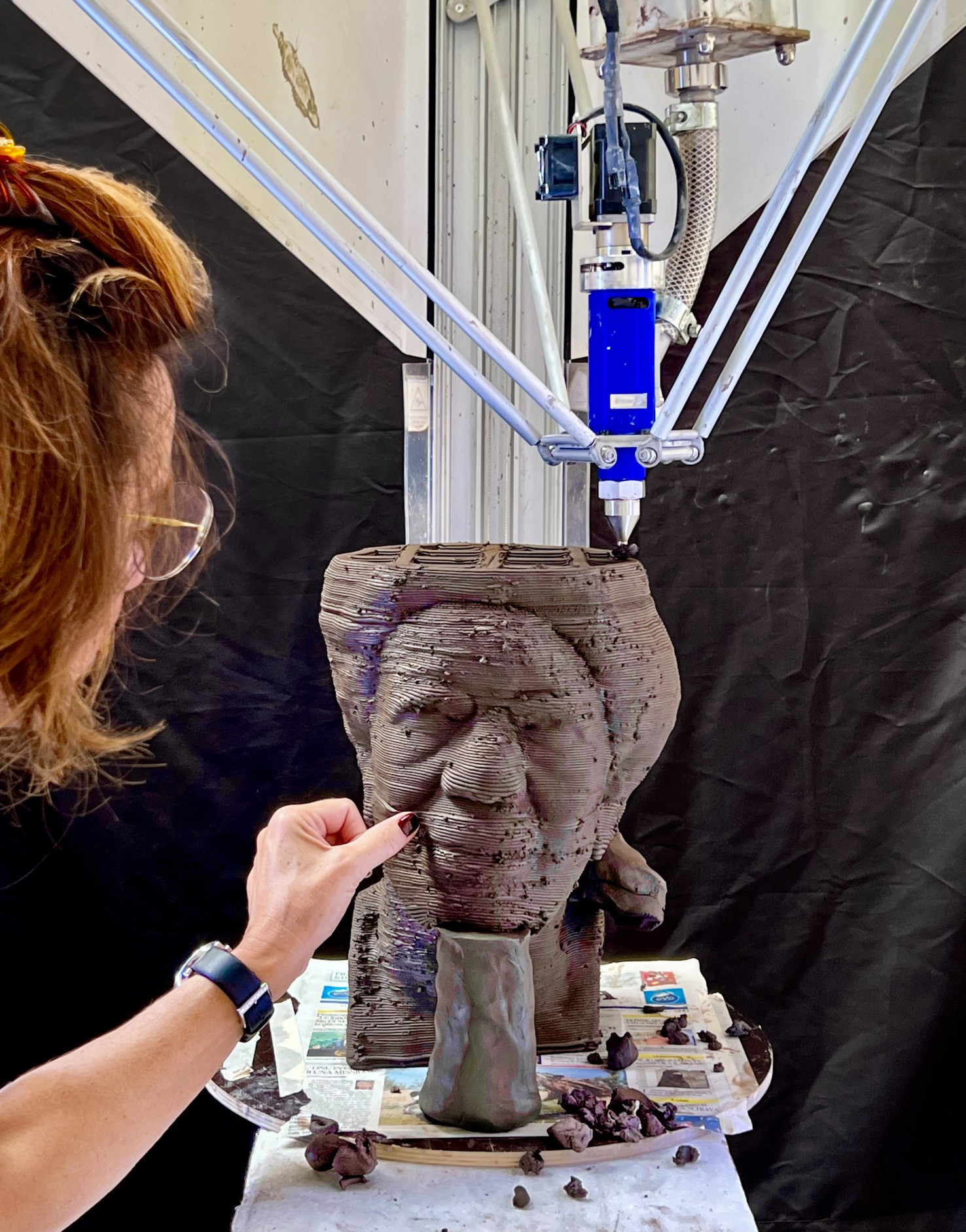 3D printing machine extruding dark grey clay as a human head with woman's hands sculpting it