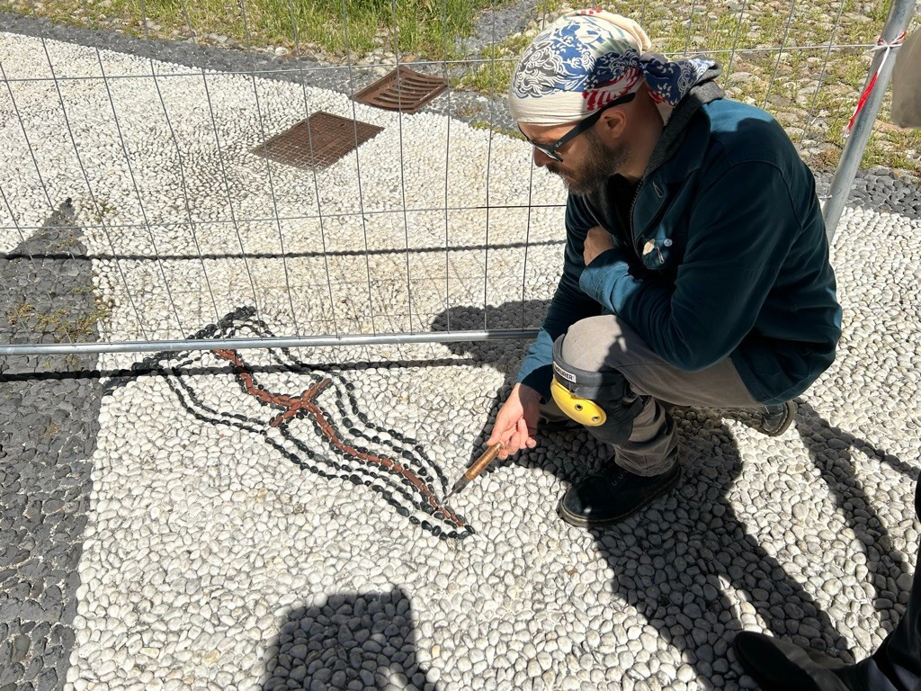 artisan pointing to his mosaic pebbles work of a flag