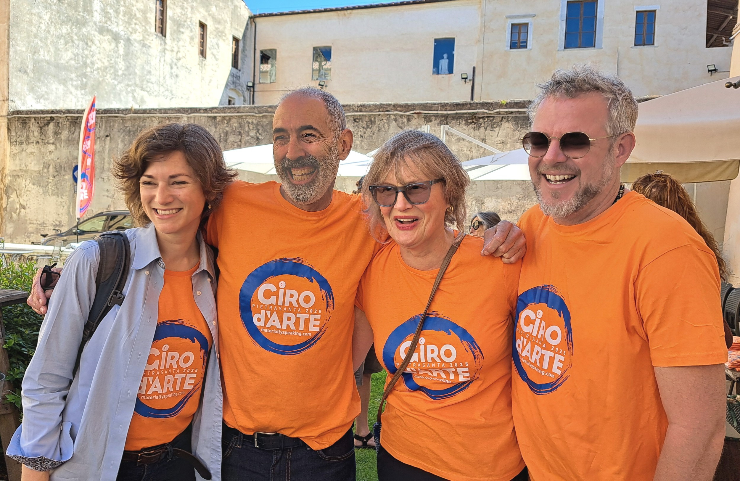 Four smiling adults wearing orange t-shirts of the 2025 Giro d'Arte in Pietrasanta, Italy