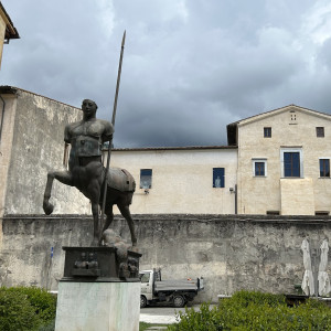 Stone buildings and stone wall with a metal disheveled statue of a fawn and a spear.