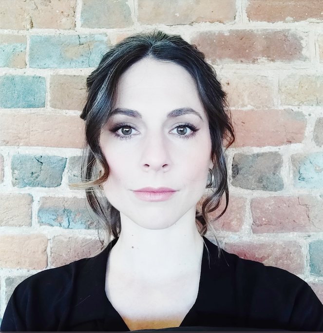 white female with brown wavy hair against a brick wall