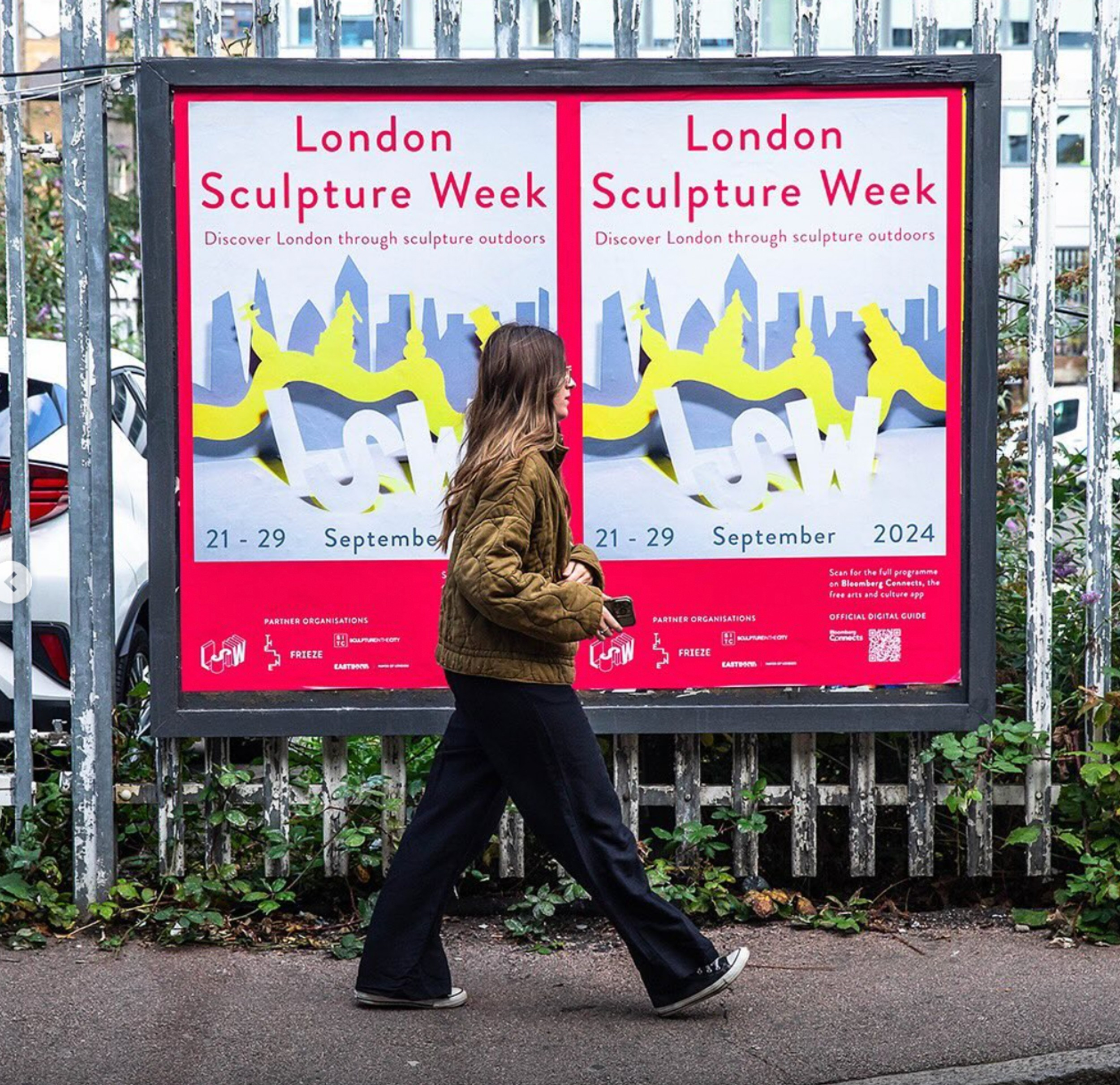 lady walking in front of a London sculpture week poster on iron railed fence