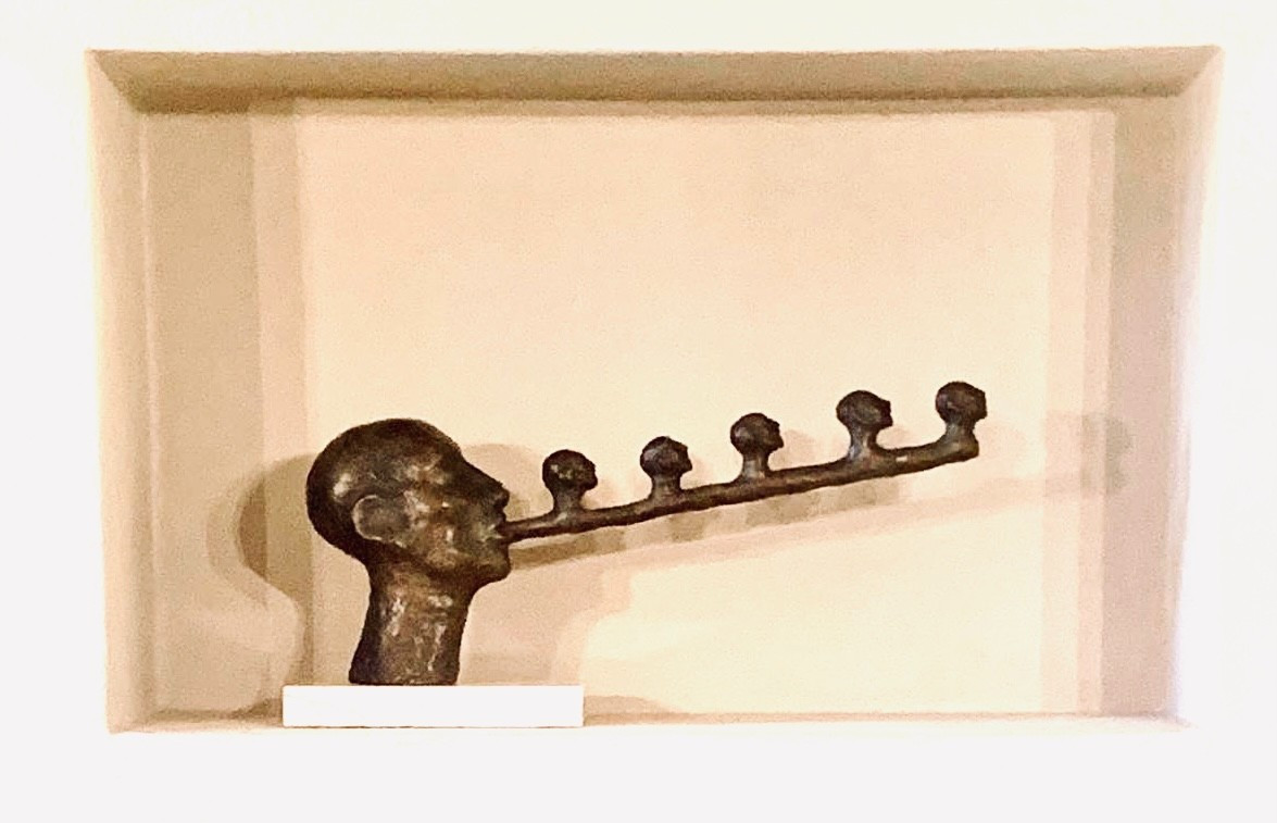 Small bronze sculpture of a head, the head's tongue extrudes and has five more heads on it.