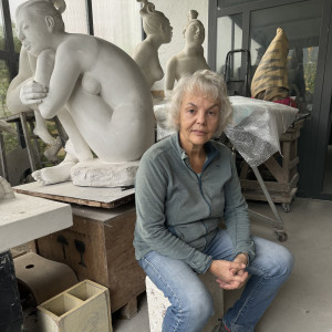 Female artist sitting on a stool among white marble figurative sculptures