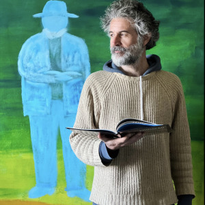 man with grey hair and beard holding and book standing in front of a painting of a blue man holding a book with a green background.