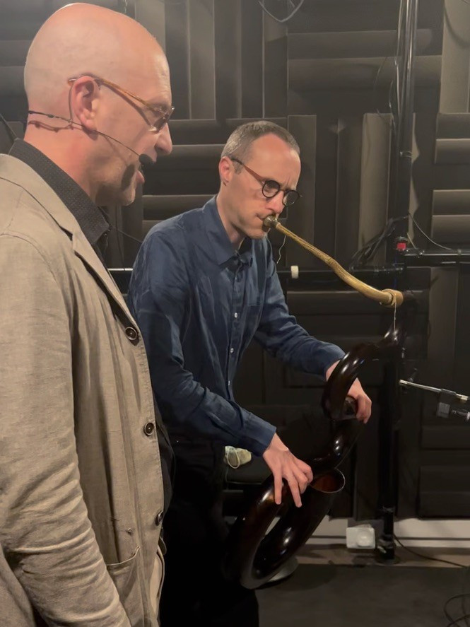 two men in audio studio, one singing and the other playing a serpent