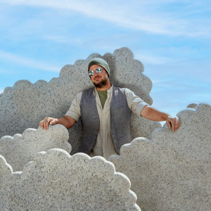 man wearing hat and sunglasses standing among cloud sculptures against a blue sky