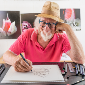 man wearing hat and glasses with white hair and beard drawing in pen in an art studio.